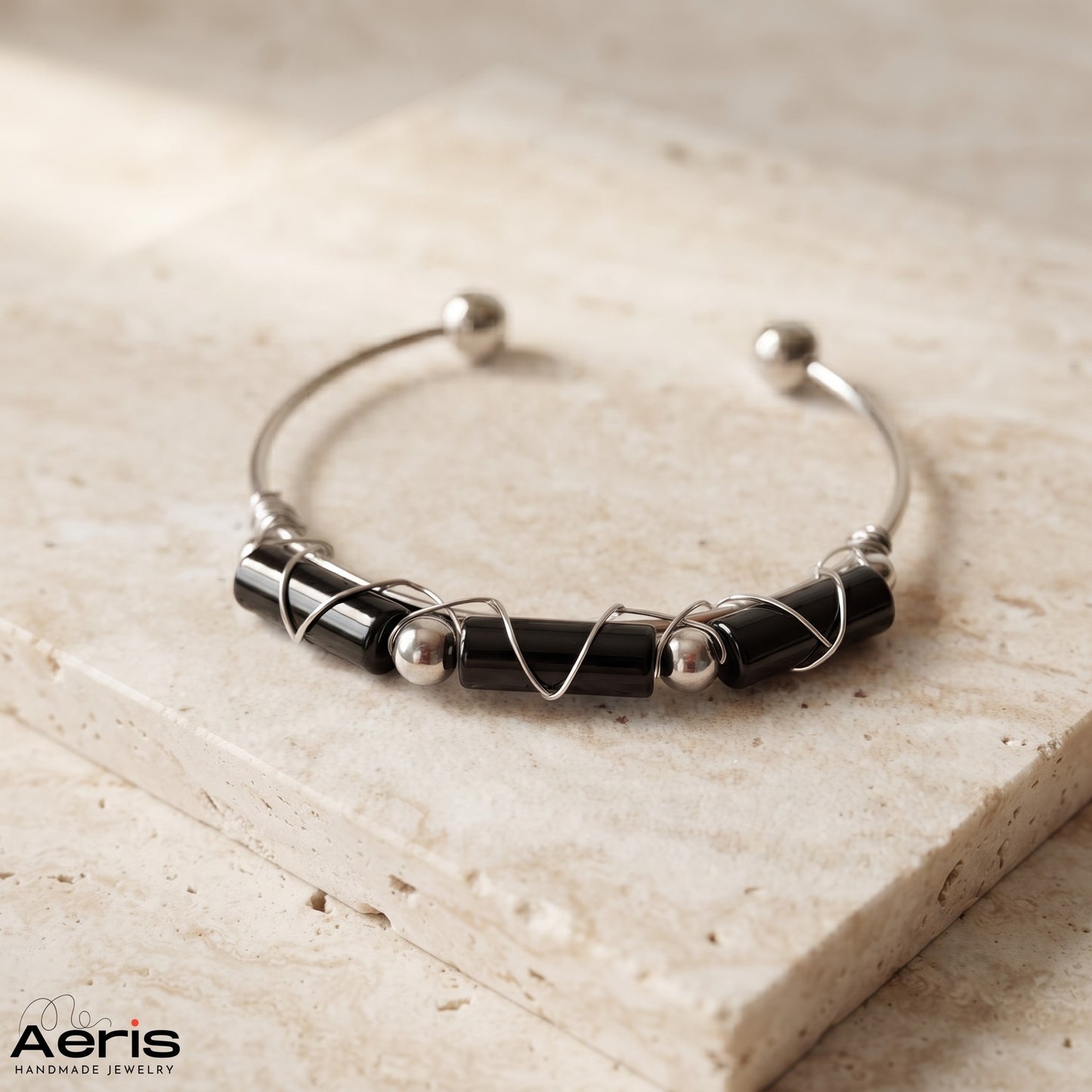 Bratara bangle cu onix tubular negru, pe o baza de inox, expusa pe o placa din travertin
