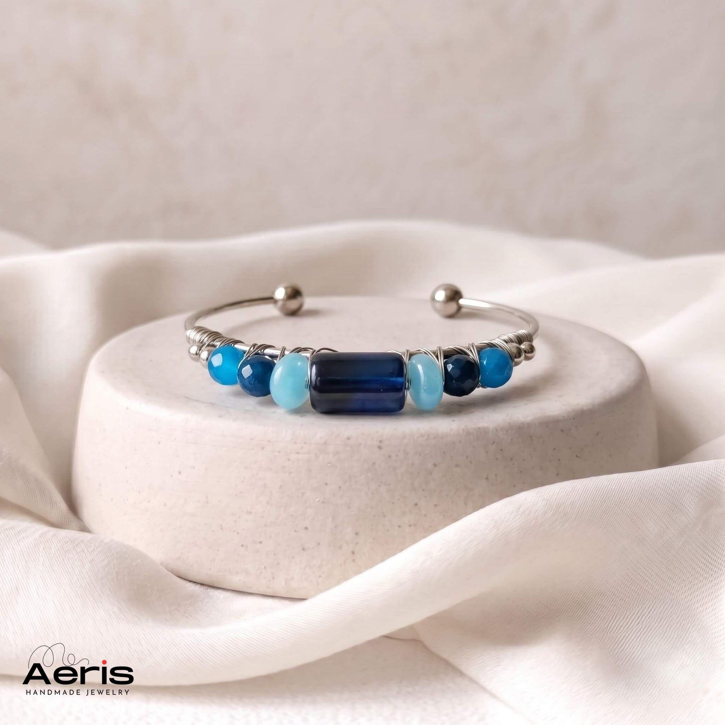 Bratara artizanala moderna cu agate albastre inserate pe o baza de inox si asezata pe un suport ceramic