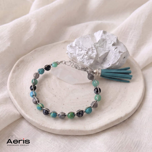Bratara handmade boho chic cu ciucure turcoaz, prezentata pe un pad ceramic, langa un cristal de cuart si o piatra cioplita