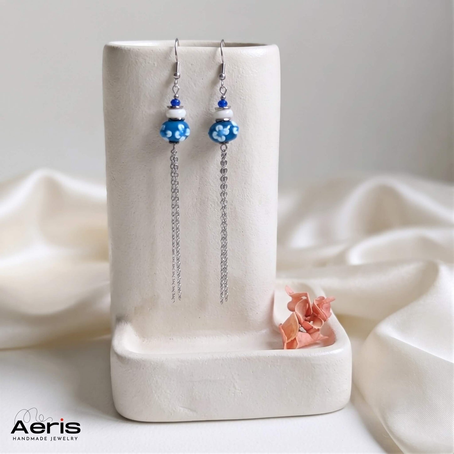 Cercei cu carlig si margele lampwork albastre cu floricele albe, prezentati pe un suport ceramic