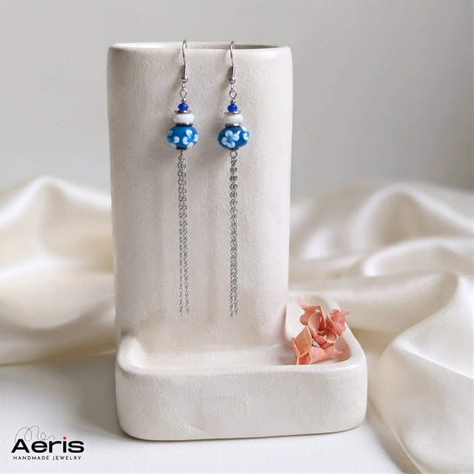 Cercei cu carlig si margele lampwork albastre cu floricele albe, prezentati pe un suport ceramic